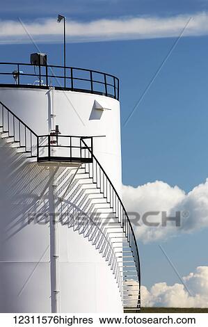 Stock Photograph - Close up of a section of a large white storage tank with black iron stairs casting a shadow on the tank and blue sky and clouds; Alberta, Canada. Fotosearch