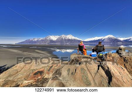 Stock Image - Couple sitting and enjoying the view of the Chugach Mountains along Turnagain Arm near Hope on the Kenai Peninsula, Southcentral Alaska, Spring, HDR. Fotosearch