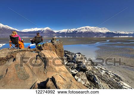 Stock Image - Couple sitting and enjoying the view of the Chugach Mountains along Turnagain Arm near Hope on the Kenai Peninsula, Southcentral Alaska, Spring, HDR. Fotosearch