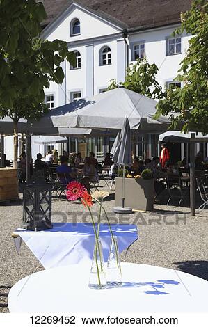 Stock Image - Customers sitting on an outdoor restaurant patio with flowers decorating a table in the foreground; Die Herreninsel im Chiemsee, Bayern, Germany. Fotosearch
