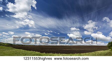 Stock Photograph - Dark soil field with green rolling hills in the background and dramatic clouds with blue sky, West of Calgary; Alberta, Canada. Fotosearch
