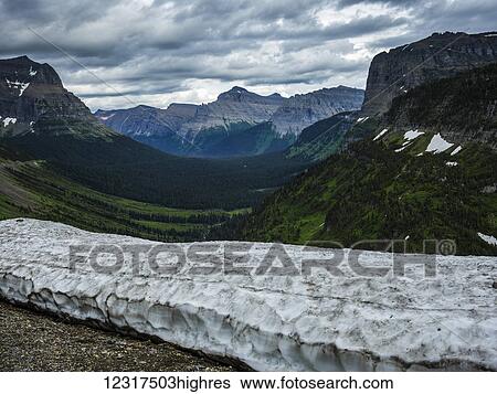 Stock Photograph - Dirty snow in a drift in the foreground with rugged cliffs and mountains in the background; Browning, Montana, United States of America. Fotosearch