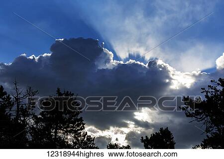 Stock Photograph - Dramatic backlit cloud covered sky with sun rays beaming through the opening; Waterton, Alberta, Canada. Fotosearch