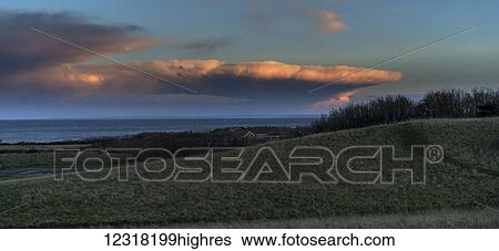 Stock Photograph - Dramatic cloud formation glowing pink at sunset over the ocean with green fields in the foreground; South Shields, Tyne and Wear, England. Fotosearch