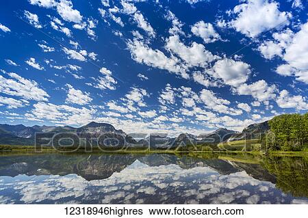 Stock Photograph - Dramatic clouds reflecting on a tranquil alpine lake with mountain range in the distance and blue sky; Waterton, Alberta, Canada. Fotosearch