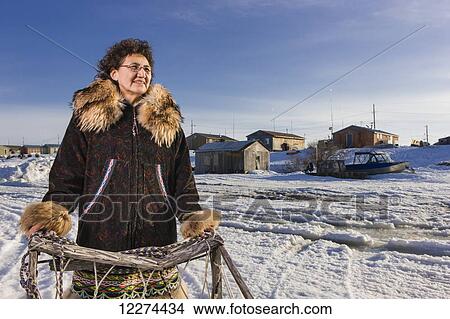 Picture - Elder Alaska native woman wearing a Kuspuk parka stands next to a wooden mushing sled, Winter, Noatak, Arctic Alaska, USA. Fotosearch