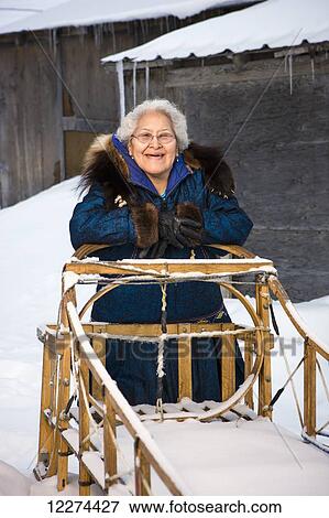 Stock Photo - Elder Alaska native woman wearing a Kuspuk parka stands next to a wooden mushing sled, Winter, Noatak, Arctic Alaska, USA. Fotosearch
