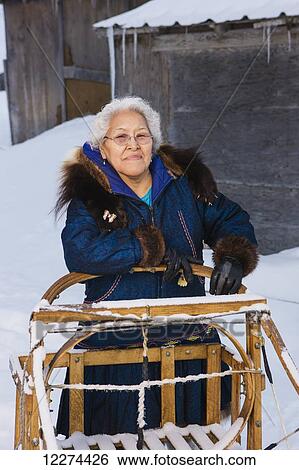 Stock Photograph - Elder Alaska native woman wearing a Kuspuk parka stands next to a wooden mushing sled, Winter, Noatak, Arctic Alaska, USA. Fotosearch