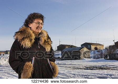 Stock Photograph - Elder Alaska native woman wearing a Kuspuk parka stands in front of village homes, Winter, Noatak, Arctic Alaska, USA. Fotosearch