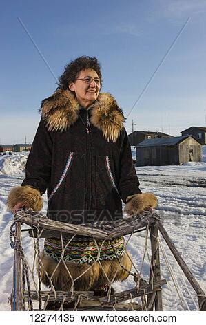 Stock Photography - Elder Alaska native woman wearing a Kuspuk parka stands next to a wooden mushing sled, Winter, Noatak, Arctic Alaska, USA. Fotosearch