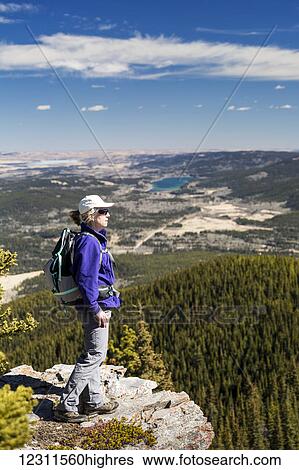 Stock Photograph - Female hiker on rocky cliff edge overlooking valley below with rolling hills, lake, blue sky and clouds, Kananaskis Country; Alberta, Canada. Fotosearch