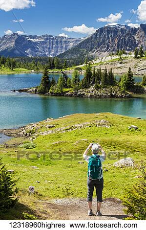 Stock Photograph - Female hiker standing on a trail taking a photo with her phone of an alpine lake with mountains, blue sky and clouds; Banff, Alberta, Canada. Fotosearch