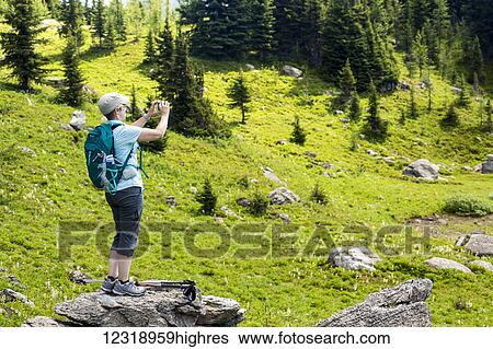Stock Photograph - Female hiker standing on top of a large rock taking a photo with her phone of an alpine meadow; Banff, Alberta, Canada. Fotosearch
