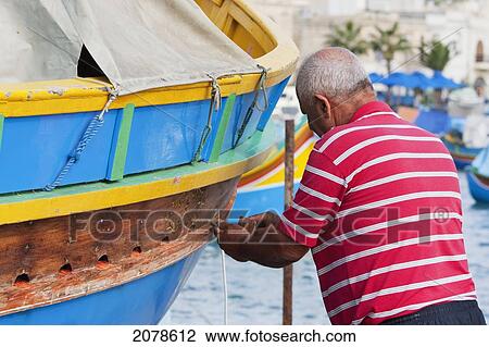 Stock Image - Fisherman Fixing His Luzzu, A Traditional Maltese Fishing Boat On The Beach, Marsaxlokk, Malta. Fotosearch
