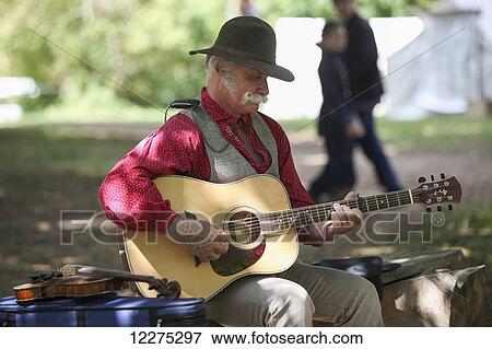 Folk musician with gray hair and a handlebar moustache playing a guitar while seated outdoors, a fiddle sitting beside him; United States of America View Large Photo Image Stock Photo - Folk musician with gray hair and a handlebar moustache playing a guitar while seated outdoors, a fiddle sitting beside him; United States of America. Fotosearch