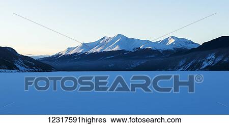 Stock Photograph - Frozen snow covered lake with snow covered mountains in the background, Liard River Hot Springs Provincial Park; British Columbia, Canada. Fotosearch