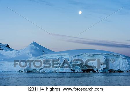 Stock Photograph - Full moon in a blue sky over the snow covered mountains and blue ocean; Antarctica. Fotosearch