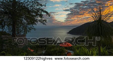 Stock Photograph - Glowing golden clouds at sunset over the pacific ocean and a silhouetted coastline and a red umbrella at a resort on the shore; Jalisco, Mexico. Fotosearch