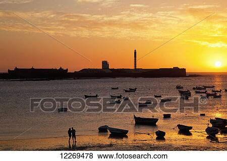 Stock Photo - Golden glowing sunset with silhouette of people on the beach and boats moored in the water; Cadiz, Andalusia, Spain. Fotosearch