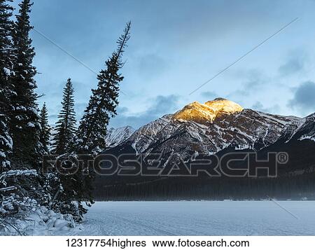 Stock Photograph - Golden sunlight on the peak of a mountain with a frozen lake in the foreground, Jasper National Park; Jasper, Alberta, Canada. Fotosearch