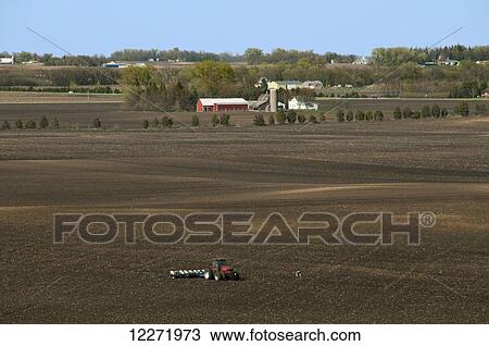 Stock Image - High expansive view of a tractor and planter in a large field planting corn in spring with farmsteads in the background, near Northrop; Minnesota, United States of America. Fotosearch
