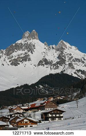 Stock Image - Hot air balloons floating high above wooden chalets and snowy mountain peaks during the ski resort's annual hot air balloon festival; Filzmoos, Austria. Fotosearch