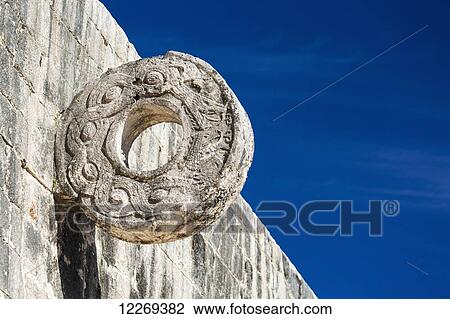 Stock Image - Large carved stone ring on side of stone wall with blue sky; Chichen Itza, Yucatan, Mexico. Fotosearch
