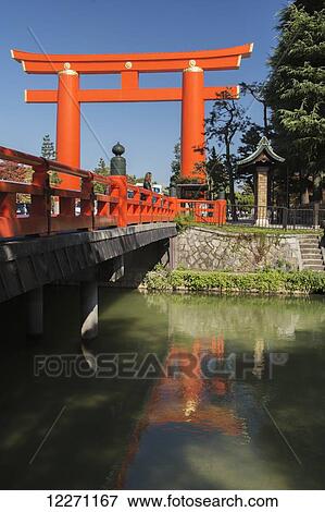 Stock Photo - Large tori gate seen from a river bank and bridge; Kyoto, Japan. Fotosearch