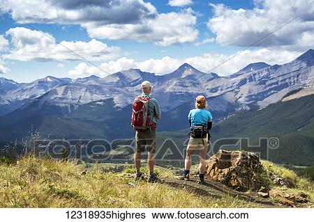 Stock Photograph - Male and female hikers standing on top of rocky hill overlooking mountain range and valley with blue sky and clouds, West of Bragg Creek; Alberta, Canada. Fotosearch