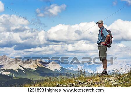 Stock Photograph - Male hiker standing on top of rocky hill overlooking mountain range and valley with blue sky and clouds, West of Bragg Creek; Alberta, Canada. Fotosearch