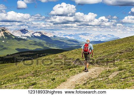 Stock Photograph - Male hiker walking along a hillside trail overlooking mountain range and valley with blue sky and cloud, West of Bragg Creek; Alberta, Canada. Fotosearch