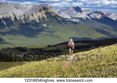 Stock Photograph - Male hiker walking along a hillside trail overlooking mountain range and valley with a cloudy sky, West of Bragg Creek; Alberta, Canada. Fotosearch