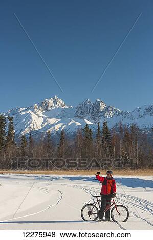 Stock Photo - Man bicycling on frozen Rabbit Creek in the Palmer Haystack Game Refuge takes a selfie with Twin Peaks in the background, Southcentral Alaska, winter. Fotosearch