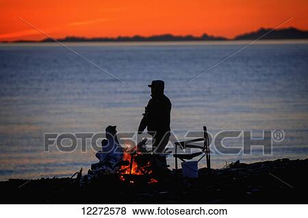 Stock Photo - Man enjoying a campfire on the beach during deep red sunset, Homer Spit, Kenai Peninsula, Southcentral Alaska, Spring. Fotosearch