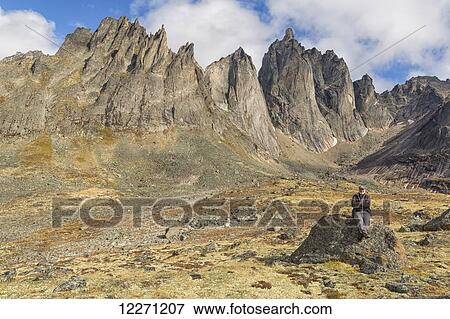 Stock Photo - Man sitting on a rock underneath the jagged granite peaks of Tombstone Territorial Park; Yukon, Canada . Fotosearch