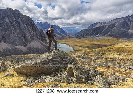 Stock Photo - Man standing on a rock overlooking the granite peaks of Tombstone Territorial Park in autumn; Yukon, Canada . Fotosearch