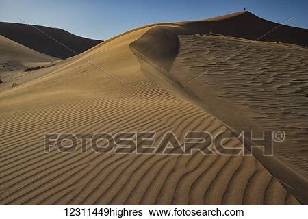 Stock Photograph - Man standing on top of a sand dune in the very strong winds buffeting the sand dunes in Sossusvlei; Namibia. Fotosearch