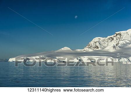 Stock Photograph - Moonset in a blue sky over the snow covered mountains and blue ocean; Antarctica. Fotosearch