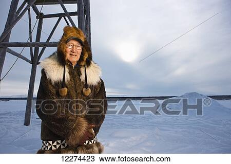 Stock Image - Native Alaskan elder in a beaver fur parka stands in front of a church bell during winter, Noatak, Arctic Alaska, USA. Fotosearch
