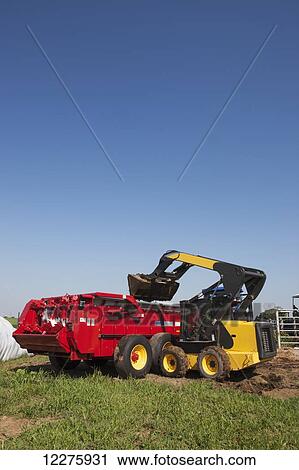 Stock Image - New Holland L180 skid steer loader loading manure into New Holland M2090 attached to T7060 tractor; New Holland, Pennsylvania, United States of America. Fotosearch
