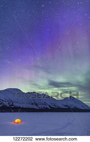 Stock Image - Nighttime view of a lit tent on a snow covered Trail Lake with the moon, stars, and Northern Lights overhead, Moose Pass, Kenai Peninsula, Southcentral Alaska. Fotosearch