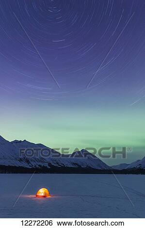 Stock Photograph - Nighttime view of a lit tent on a snow covered Trail Lake with the moon, stars, and Northern Lights overhead, Moose Pass, Kenai Peninsula, Southcentral Alaska. Fotosearch