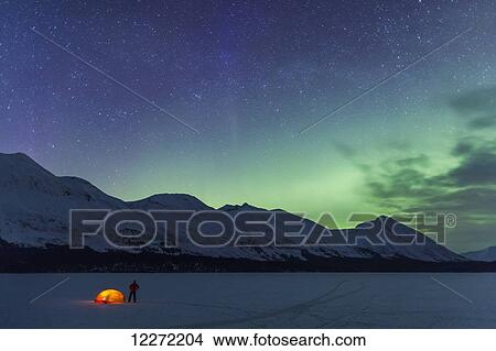 Picture - Nighttime view of a man standing next to a lit tent on a snow covered Trail Lake with the moon, stars, and planets overhead, Moose Pass, Kenai Peninsula, Southcentral Alaska. Fotosearch
