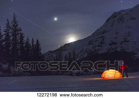 Stock Photo - Nighttime view of a man standing next to a lit tent on a snow covered Trail Lake with the moon, stars, and planets overhead, Moose Pass, Kenai Peninsula, Southcentral Alaska. Fotosearch