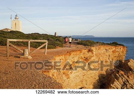Pedestrians walk on a path on a cliff ledge with lighthouse on the coast; Conil de la Frontera, Andalusia, Spain View Large Photo Image Stock Image - Pedestrians walk on a path on a cliff ledge with lighthouse on the coast; Conil de la Frontera, Andalusia, Spain. Fotosearch