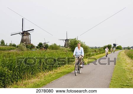 Stock Image - People Cycling By Windmills, Built Around 1740 Along Nieuwe Waterschap To Drain The Excess Water From The Alblasserwaard Polders, Kinderdijk, Netherlands. Fotosearch