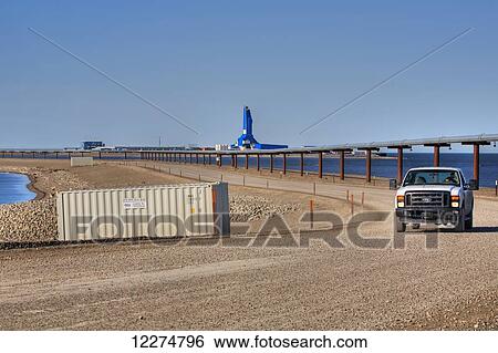 Stock Photograph - Pickup truck driving on gravel road in Prudhoe Bay Oilfield with the Liberty Oil Rig and pipeline in the background, Arctic Alaska, Summer. Fotosearch