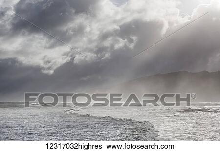 Stock Photograph - Rays break through stormy clouds over breaking waves; Greymouth, South Island, New Zealand. Fotosearch
