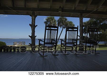 Stock Photography - Rocking chairs on a covered porch overlooking a grassy lawn and palm trees with the coastal water in the background; United States of America. Fotosearch