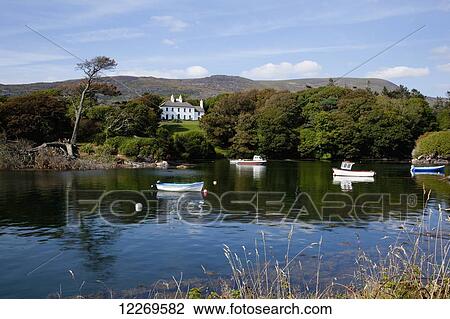 Stock Image - Rowboats moored in the tranquil water with a large white house on the shore, near Castlecove; County Kerry, Ireland. Fotosearch